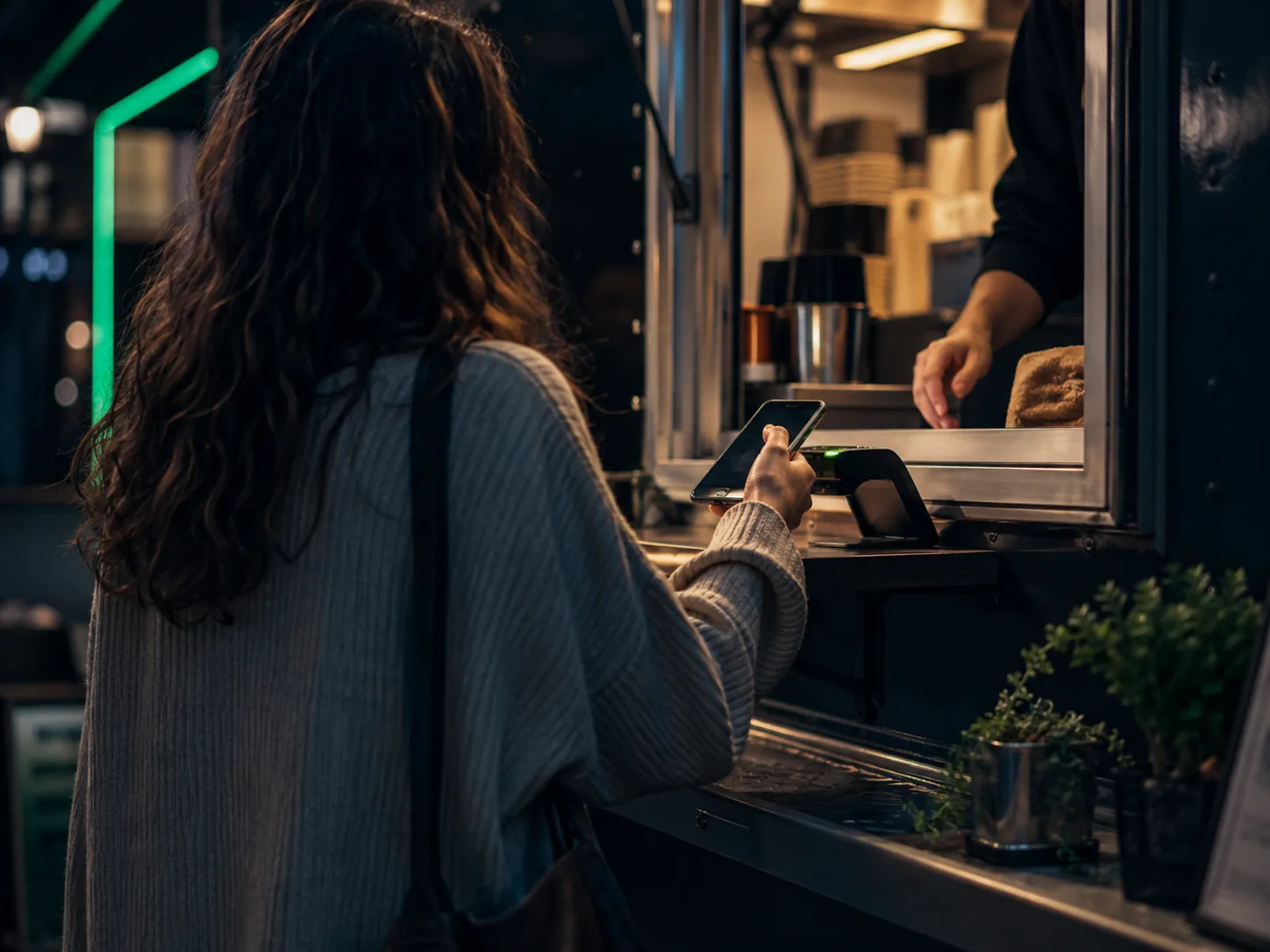 Person paying with a phone at a food-truck window in the evening with a green neon glow