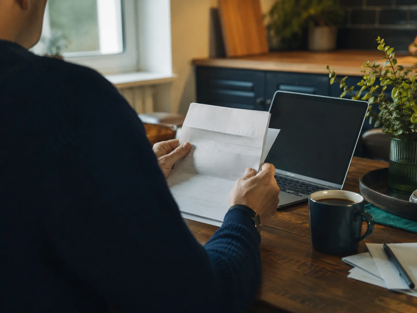 Person at a kitchen table reviewing paper bills beside a laptop and coffee in warm morning light